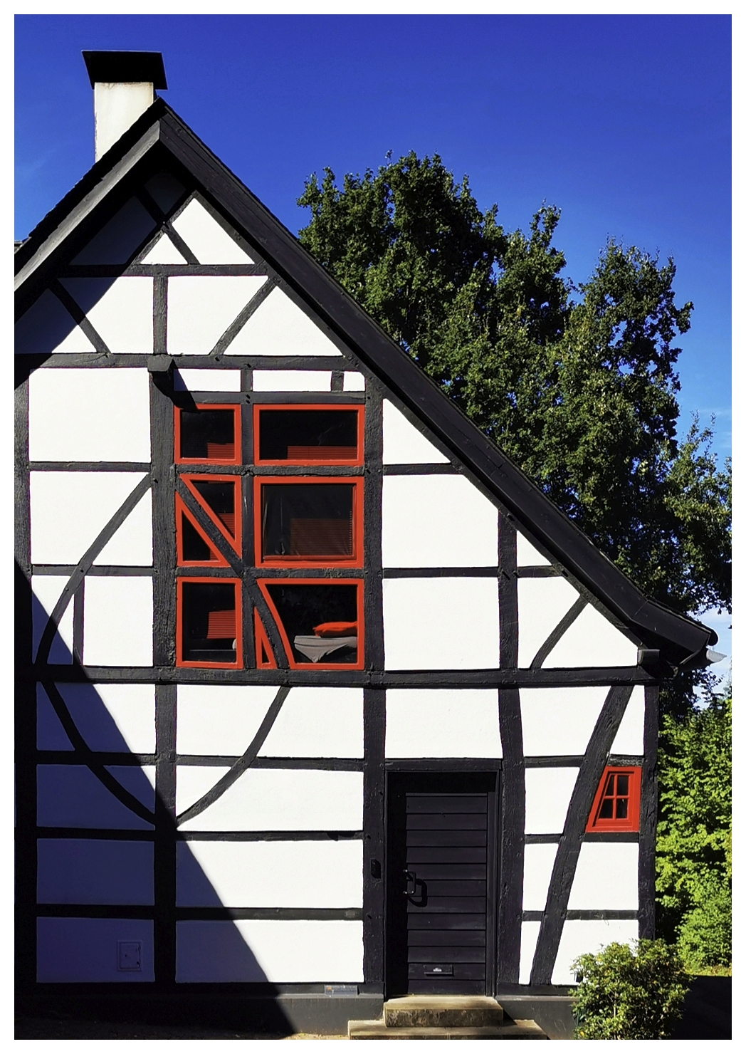 Fachwerkhaus mit weißer Fassade und schwarzen Balken, rote Fensterrahmen und schwarze Tür. Die geometrischen Muster der Balken sind deutlich sichtbar. Im Hintergrund steht ein grüner Baum vor blauem Himmel. Der Schatten eines anderen Gebäudes fällt auf die linke Seite der Hauswand.