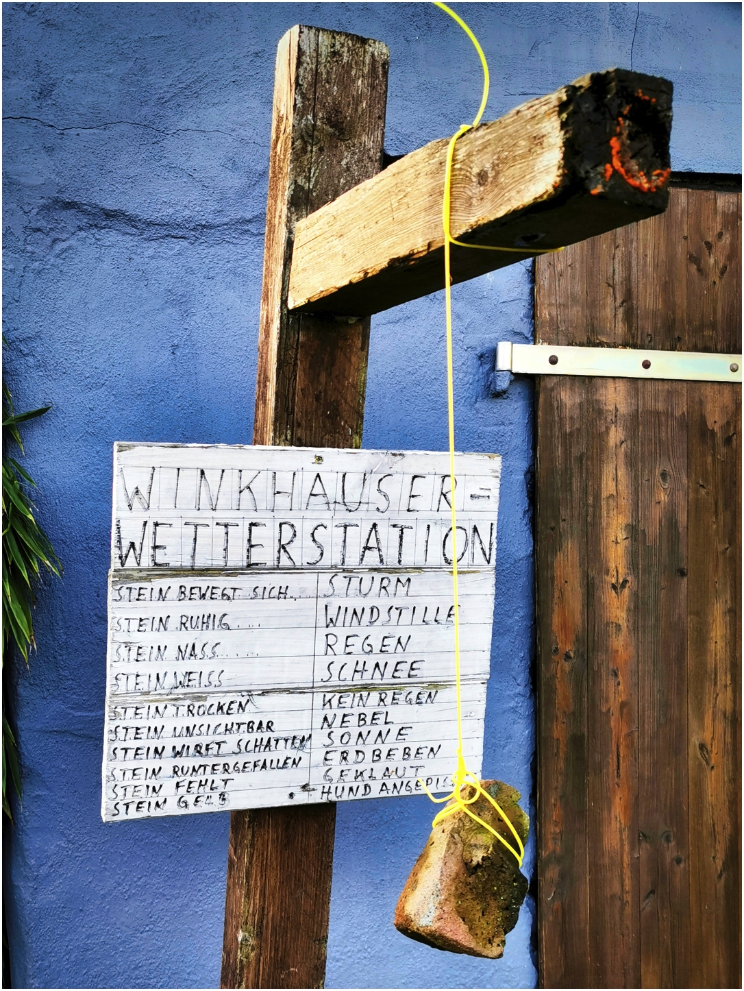 Im Mittelpunkt steht ein hölzernes Schild mit der Aufschrift 'Winkhäuser Wetterstation', das an einem Holzbalken befestigt ist. Darunter sind in tabellarischer Form verschiedene Wetterlagen und dazu passende Zustände eines Steins aufgelistet, etwa 'Stein bewegt sich – Sturm', 'Stein nass – Regen', 'Stein trocken – kein Regen'. Am gleichen Holzbalken hängt an einer gelben Schnur ein faustgroßer Stein frei vor  dem Schild. Der Hintergrund besteht aus einer blauen Wand und einer Holztür. Das Schild und der Stein sind zentrale Elemente und illustrieren humorvoll eine sogenannte Wetterstation, die das Wetter anhand des Zustands des Steins beschreibt.