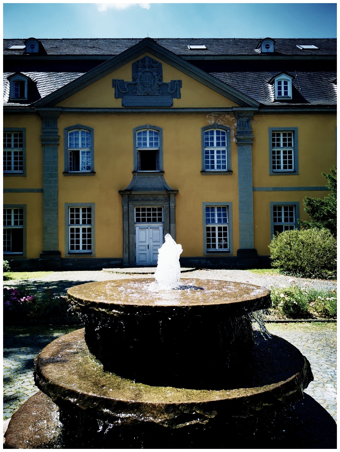 Im Vordergrund steht ein mehrstufiger Brunnen mit sprudelndem Wasserstrahl. Dahinter befindet sich ein großes, gelbes Gebäude mit symmetrischer Fassade, weißen Fensterrahmen und einem zentralen Eingang mit steinernem Rahmen. Das Dach ist dunkel und mit Schiefer gedeckt. Über dem Eingang ist ein Wappen oder Relief zu erkennen. Die Szene ist von Tageslicht beleuchtet.