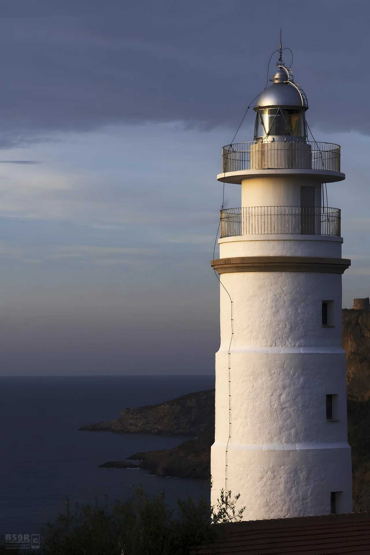 Das Foto im Portraitformat zeigt am rechten Bildrand und die Bildbreite zur Hälfte ausfüllend einen strahlend weißen Leuchtturm, welcher auf einer Klippe hoch über dem Meer steht. Die linke Bildhälfte zeigt den felsigen Küstenstreifen und gibt den Blick aufs Meer frei. Der Himmel ist wolkenlos, es ist kurz vor Einbruch der Dämmerung.



The photo in portrait format shows a gleaming white lighthouse standing on a cliff high above the sea on the right-hand edge of the picture, filling half of its width. The left half of the picture shows the rocky coastline and offers a view of the sea. The sky is cloudless, it is just before dusk.
