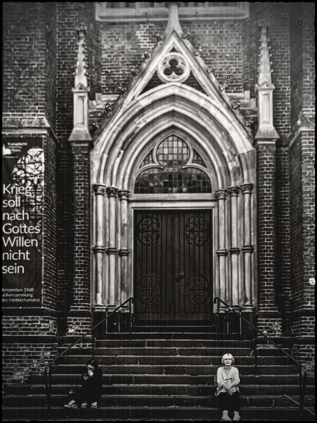 Zentral im Bild steht ein großes, spitzbogiges Kirchenportal mit reich verziertem Steinrahmen und schweren Holztüren. Die Fassade besteht aus Backstein, und über dem Portal befindet sich ein rundes Maßwerkfenster. Links neben dem Eingang hängt ein Banner mit dem Text 'Krieg soll nach Gottes Willen nicht sein'. Auf den Treppenstufen vor dem Portal sitzen zwei Personen, eine links und eine rechts, beide mit neutralem Gesichtsausdruck. Die Aufnahme ist schwarz-weiß und betont die Struktur der Backsteinmauer und die Details der Architektur.
