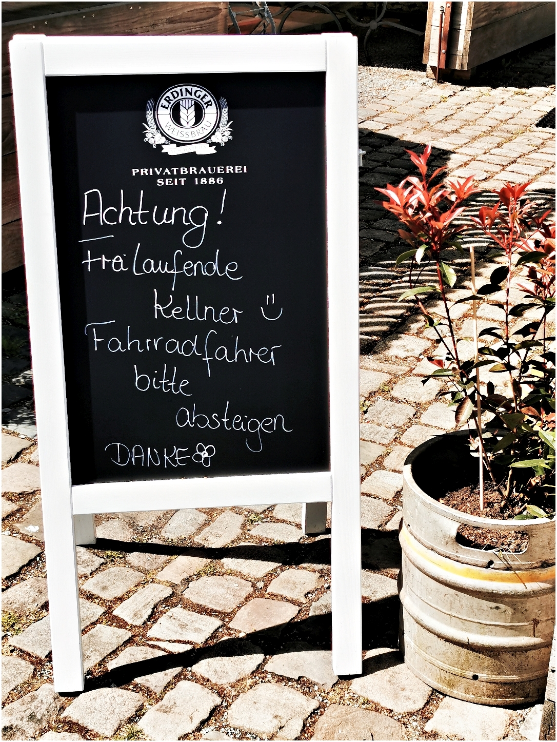 Im Vordergrund steht eine schwarze Tafel mit weißem Rahmen auf dem Kopfsteinpflaster. Die Tafel trägt einen handgeschriebenen Hinweis in weißer Kreide, der Text enthält eine 'Warnung vor freilaufenden Kellnern' und formuliert die Bitte an Fahrradfahrer, abzusteigen. Links oben ist das Logo der Erdinger Privatbrauerei zu sehen. Rechts neben der Tafel steht ein bepflanzter Bierfass-Blumenkübel mit einem kleinen roten Strauch. Im Hintergrund sind Holzelemente und Teile einer Terrasse erkennbar.