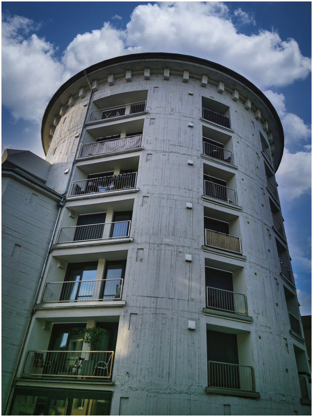 Ein runder, mehrstöckiger ehemaliger Turm- oder Hochbunker mit Betonfassade und nachträglich eingeschnitten Balkonen steht im Vordergrund der Fotografie. Der Bunker wurde einer neuen Nutzung als Wohnhaus zugeführt, da sein Abriss aufgrund der massiven Stahlbetonwände zu teuer gewesen wäre. Die Fassade zeigt vertikale Linien und kleine quadratische Elemente. Der obere Bereich ist mit einer markanten, zylindrischen Form gestaltet. Der Himmel im Hintergrund ist blau und von weißen Wolken bedeckt.