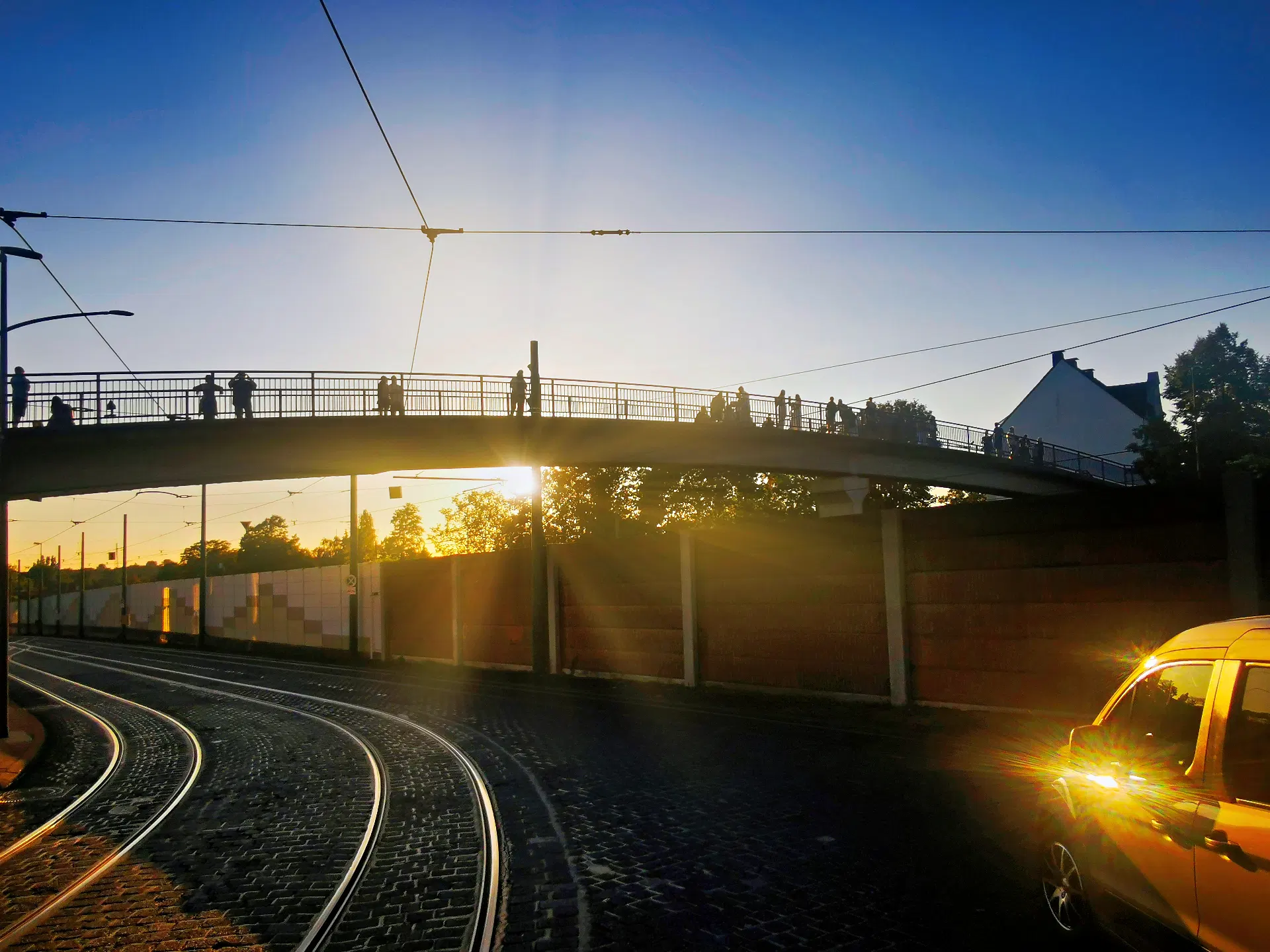Eine Brücke mit Geländer verläuft quer durch das Bild und hebt sich als Silhouette gegen einen klaren Himmel ab. Die Sonne steht tief und erzeugt einen intensiven Lichtschein, der die Konturen der Brücke und die Umrisse von Menschen darauf betont, die zur Himmelsbeobachtung auf der Brücke stehen. Im Vordergrund sind Straßenbahnschienen und ein hell beleuchtetes Fahrzeug zu sehen. Bäume und Gebäude bilden den Hintergrund.