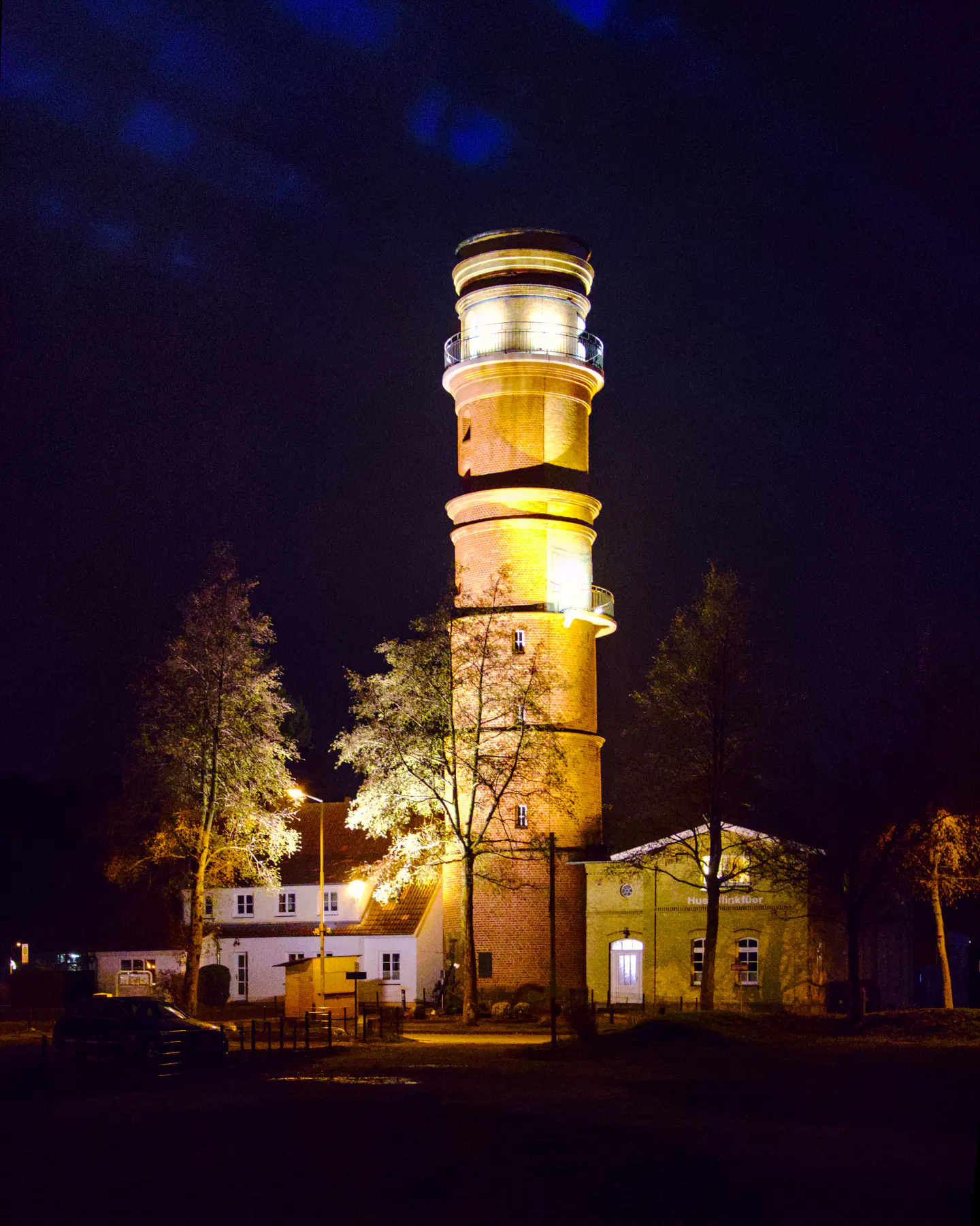 Mitten in der Nacht erscheint der alte Leuchtturm von Travemünde angestrahlt von hellen Lichtern. Die beiden Nebengebäude des Leuchtturms und auch die Bäume vor dem Leuchtturm stehen ebenfalls im Licht.