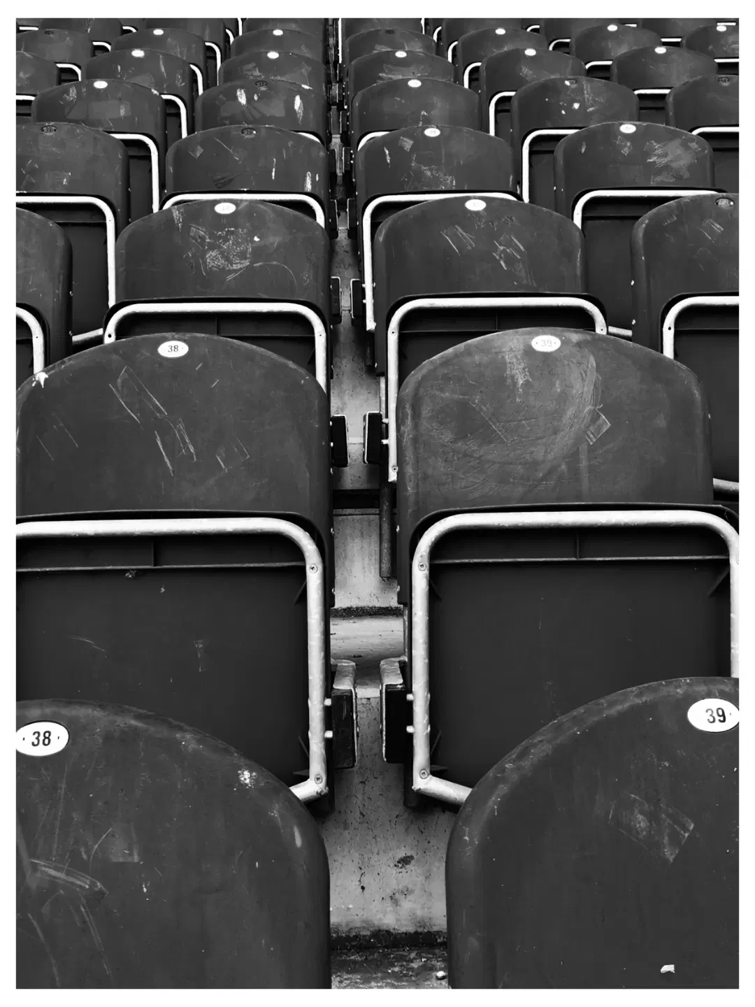 Reihen von leeren, dunkel gefärbten Stadionsitzen mit sichtbaren Gebrauchsspuren und Kratzern. Die Sitze sind durch Metallbügel voneinander getrennt. Im unteren Bereich sind runde Plaketten mit Zahlen oder Platznummern zu erkennen.
