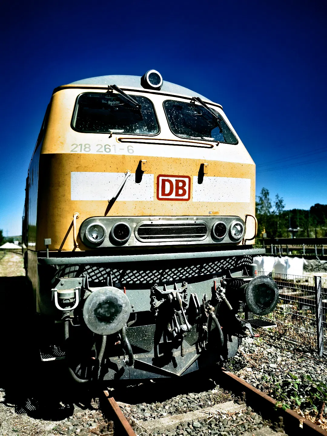 Im Vordergrund steht eine gelbe Diesellokomotive der Baureihe 218 mit der Nummer '218 261-6'. Das DB-Logo ist mittig auf der Front angebracht. Die Lokomotive befindet sich auf einem Gleis im Freien, umgeben von Kies und einem Zaun. Der Himmel ist klar und tiefblau, im Hintergrund sind Bäume und weitere Gleisanlagen zu erkennen.