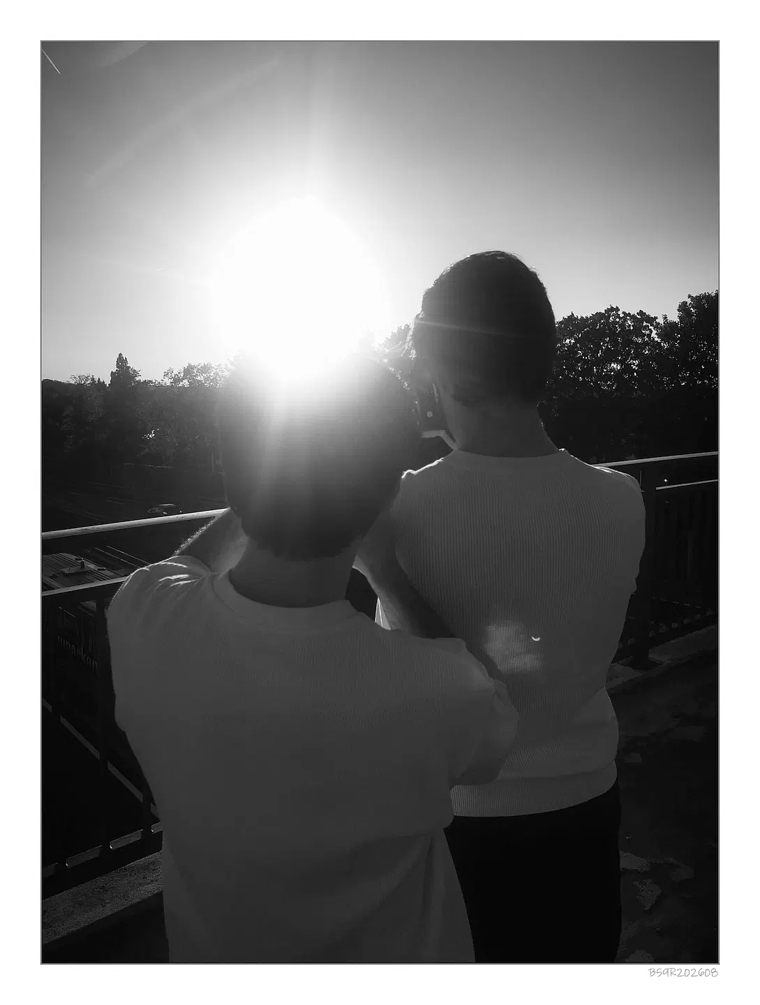 Zwei Personen stehen nebeneinander auf einer Brücke und blicken in Richtung der untergehenden Sonne in Erwartung einer Sonnenfinsternis. Die Sonne steht bereits tief am Himmel und blendet stark, sodass Details im Hintergrund nur schemenhaft erkennbar sind. Beide tragen helle Oberteile, das Licht wirft lange Schatten und betont die Konturen der Silhouetten. Im Hintergrund sind das Brückengeländer und weiter entfernt Bäume sichtbar.