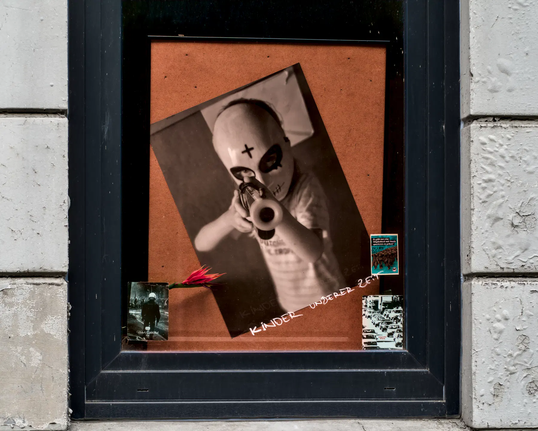 Eine große Fotografie in einem Fenster zeigt einen Jungen von vielleicht 10 Jahren mit einer Maske, die das ganze Gesicht bedeckt. Auf der Stirn prangt ein Kreuz. Der Junge hält ein Spielzeuggewehr in den Händen, mit dem er direkt auf den Fotografen zielt. Unter dem Bild steht handschrftlich: "Kinder unserer Zeit"
.
A large photograph in a window shows a boy of perhaps 10 years old wearing a mask that covers his entire face. A cross is prominently displayed on his forehead. The boy is holding a toy gun, which he is pointing directly at the photographer. Beneath the picture, the words ‘Children of our time’ are written by hand.
.