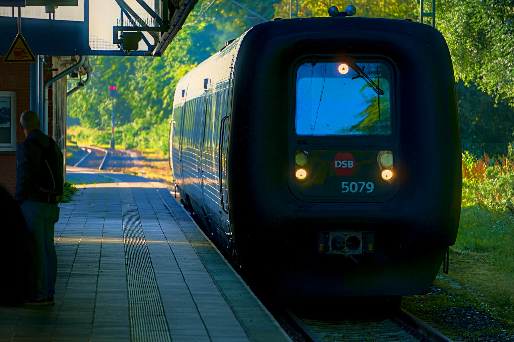 Ein Regionalzug der DSB (Danske Statsbaner) trifft am Flensburger Bahnhof ein. Links der Bahnsteig, auf dem ein einzelner Mensch dem Zug den Rücken zuwendet, rechts auf dem Gleis der eher praktische designte Zug mit drei hellen Leuchten im Dreieck an der Front. Im Hintergrund leuchtet das Grün neben den Gleisen im Licht der Morgensonne.