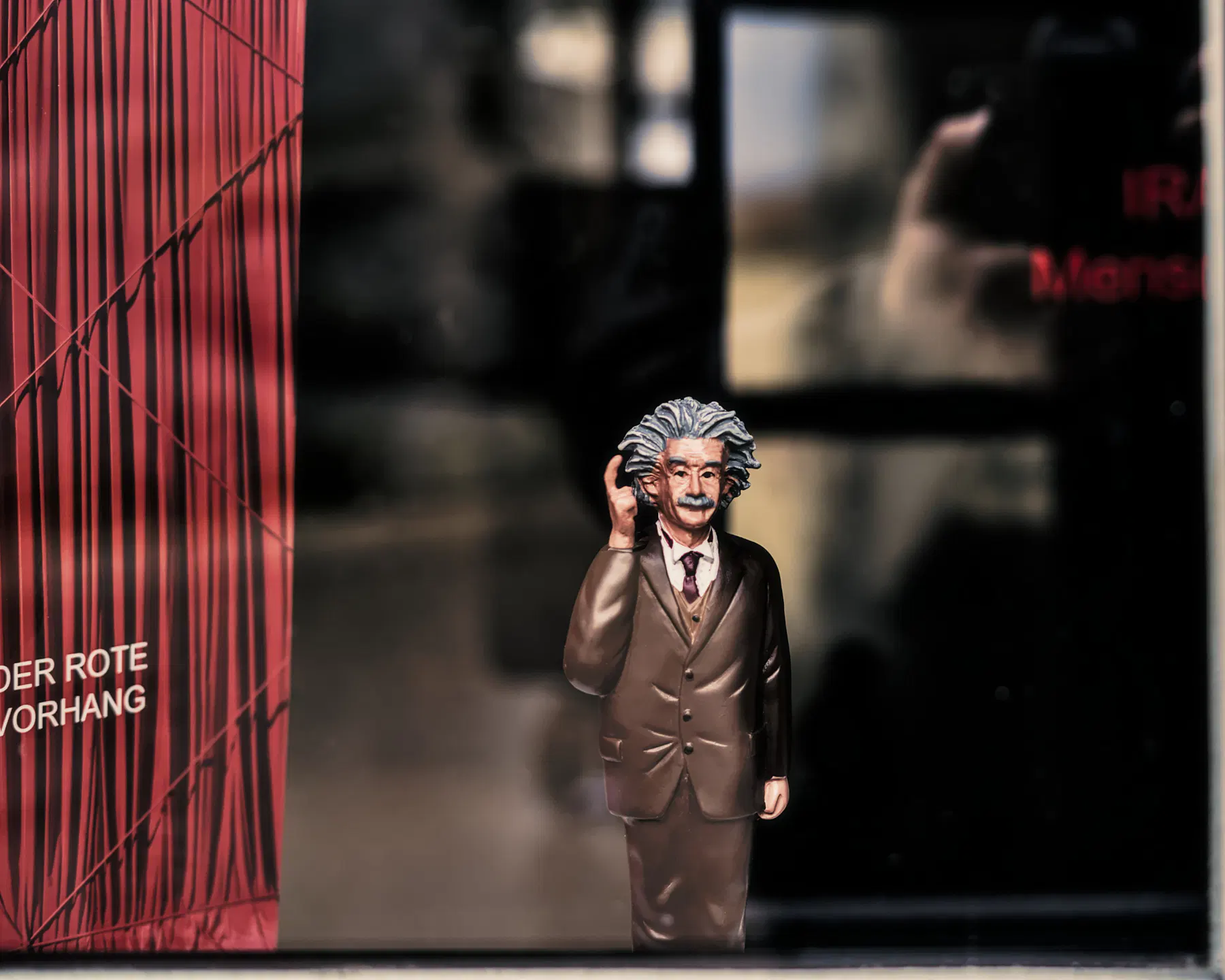 In einem Schaufenster, das links von einem roten Vorhang begrenzt wird, steht eine Figur von Albert Einstein, der sich mit dem Zeigefinger der rechten Hand an die Stirn tippt: Ihr spinnt doch!