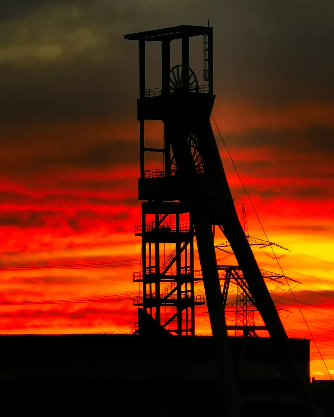 Eine markante Silhouette eines Förderturms steht vor einem dramatisch orange-roten Himmel bei Sonnenuntergang, der den Eindruck erweckt, als stünde der Himmel in Flammen. Die Struktur zeigt mehrere Räder und Streben, daneben verlaufen Stromleitungen. Die dunklen Umrisse heben sich deutlich vom leuchtenden Hintergrund ab.