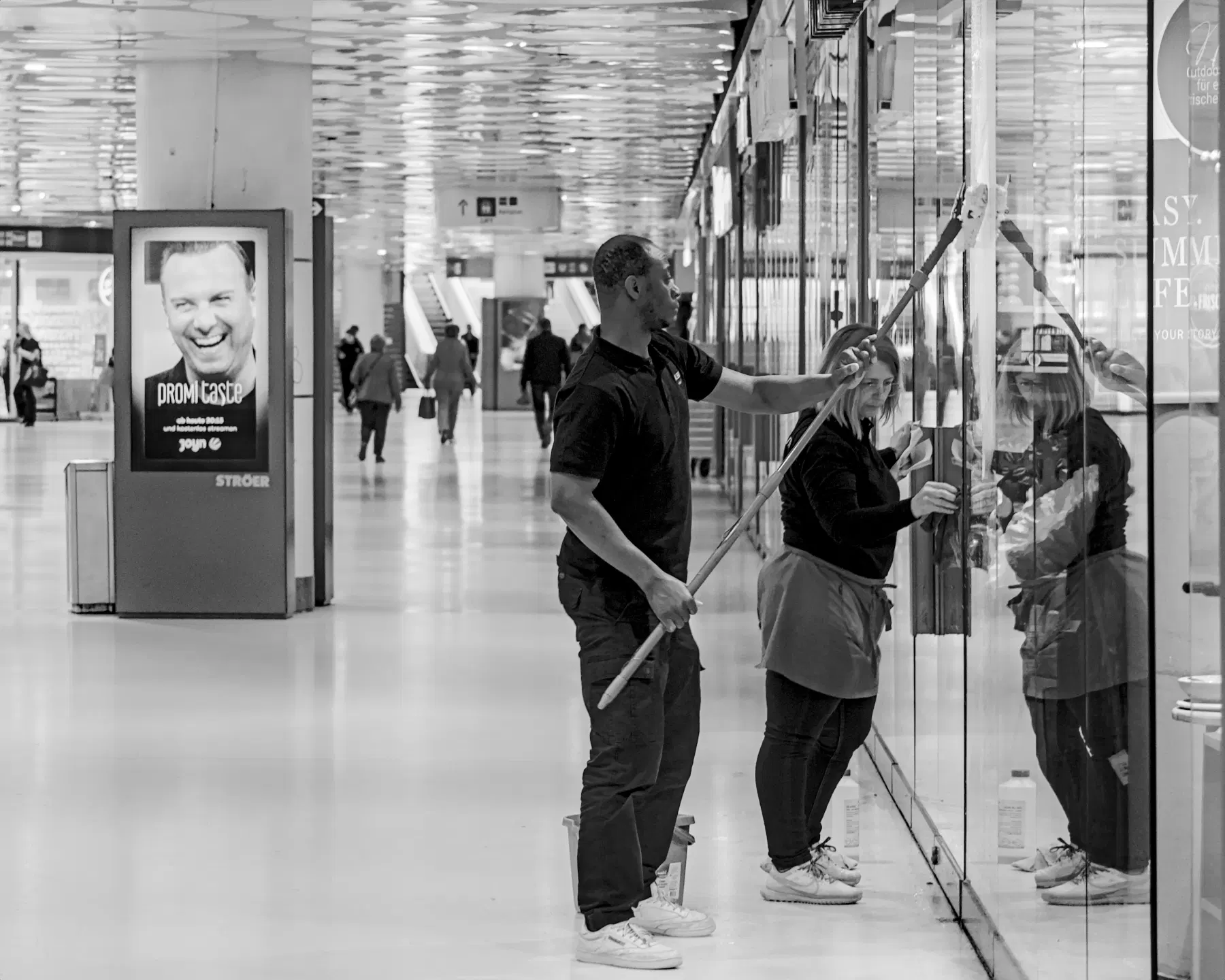 In einer hellen Einkaufspassage voller Menschen und Werbetafeln stehen zwei in schwarz gekleidete Menschen, die die Glasfront eines Geschäftes mit einem Wischer und einem Lappen reinigen. 
. 
In a bright shopping arcade full of people and advertising billboards, two people dressed in black are cleaning the glass front of a shop with a windscreen wiper and a cleaning cloth.
.