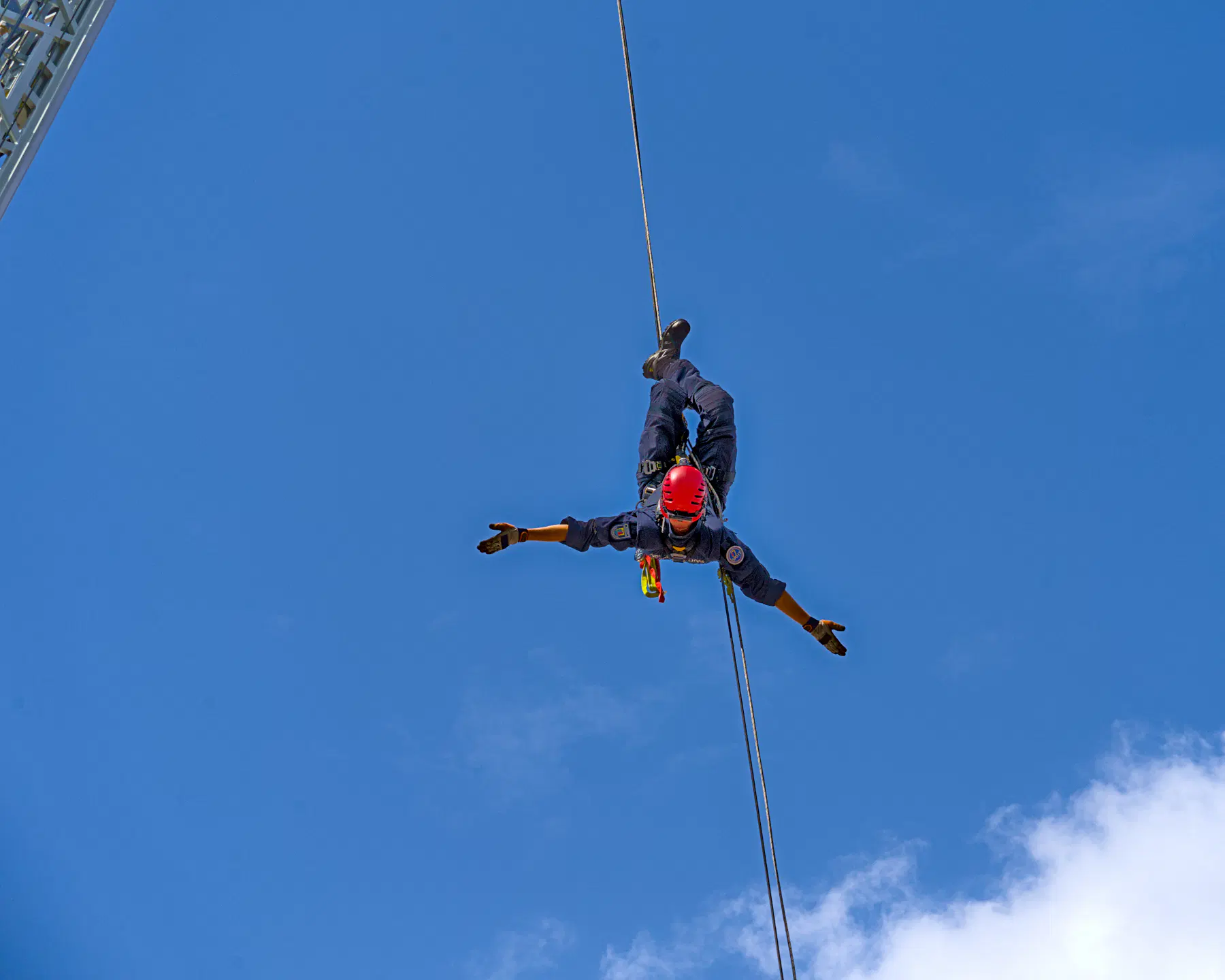 Bei einer Vorführung am Blaulichttag 2026 hängt ein Feuerwehrmann kopfüber am Seil eines Krans. Der Kran ist links oben in der Ecke gerade noch zu erkennen, rechts unten wird der Kletterer von einer weißen Wolke begrüßt. Der Rest des Himmels ist blau.