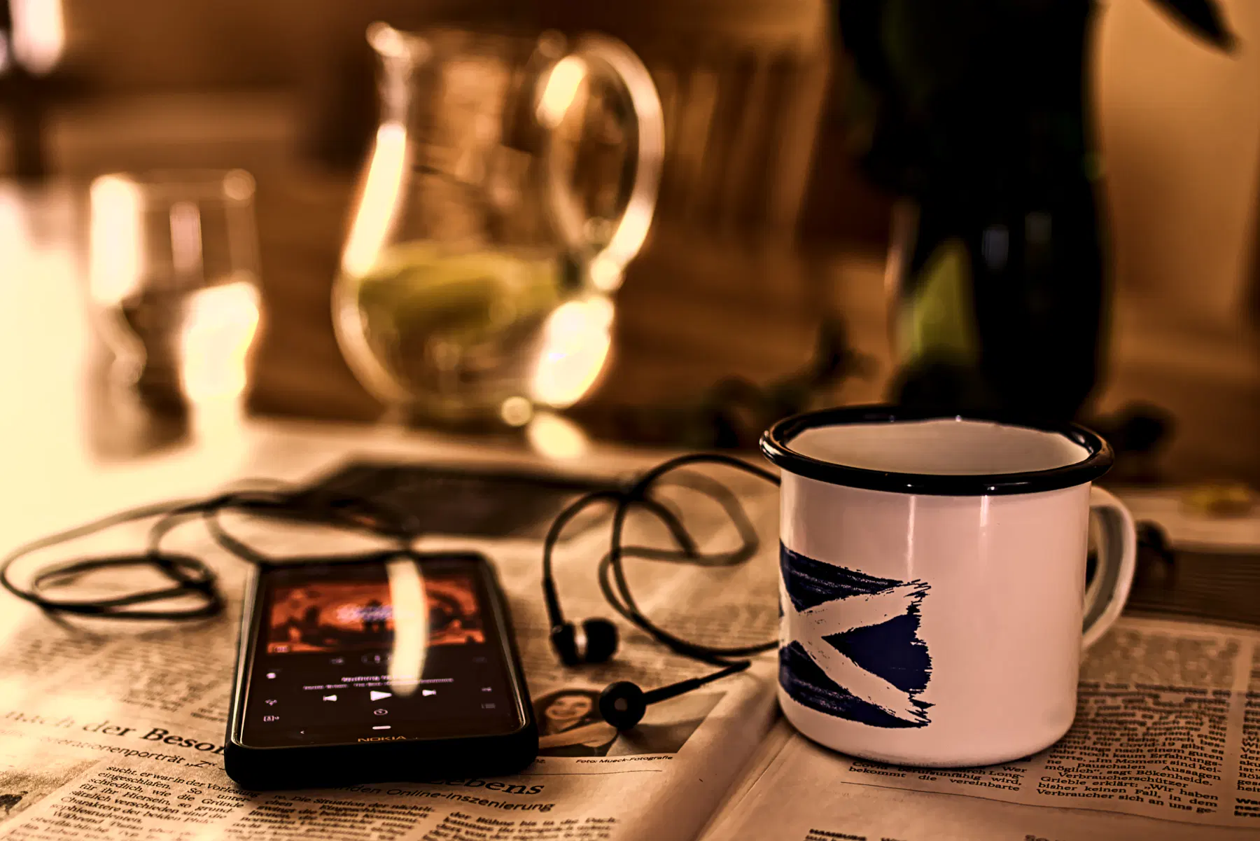 Eine Zeitung liegt aufgeschlagen auf einem Tisch, darauf ein Smartphone mit Kopfhörern und eine Emaille-Tasse mit der schottischen Flagge darauf. Im Hintergrund eine Karaffe mit selbstgemachter Limonade und ein halb gefülltes Glas.