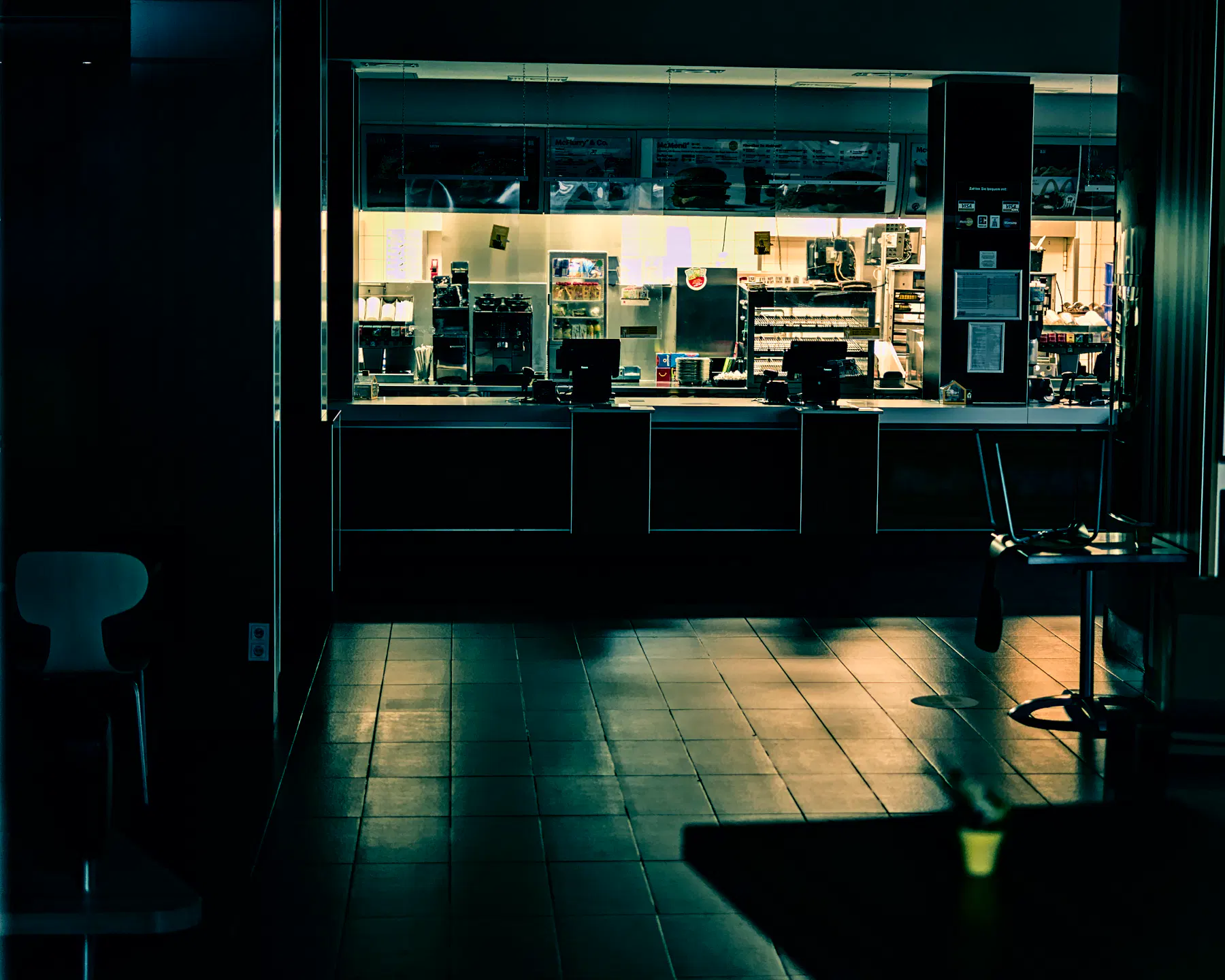 Ein dunkles Bild eines Fastfood-Restaurants. Der Raum für die Gäste ist leer dunkel, die Stühle und Tische sind nicht besetzt. Im Hintergrund sind Küche und Tresen beleuchtet. Das Licht spiegelt sich in den Fliesen des Gastraumes. Ein dunkles Bild eines Fastfood-Restaurants. Der Raum für die Gäste ist leer dunkel, die Stühle und Tische sind nicht besetzt. Im Hintergrund sind Küche und Tresen beleuchtet. Das Licht spiegelt sich in den Fliesen des Gastraumes.