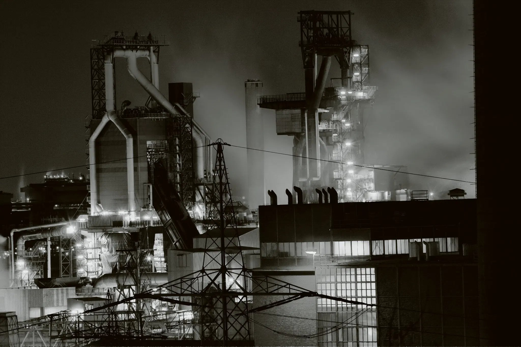 Im Vordergrund sind mehrere Gebäude mit flachen Dächern und Fensterelementen zu sehen. Dahinter erstreckt sich ein großflächiger Industriekomplex mit hohen Stahlkonstruktionen, Rohrleitungen und Schornsteinen. Die Anlage ist von zahlreichen Lichtern und Dampf umgeben, was auf einen laufenden Betrieb hindeutet. Stromleitungen und Masten durchziehen das Bild. Die Szene zeigt einen modernen Hochofen des Thyssen-Krupp Stahlwerks zu Duisburg in einer schwarz-weißen Aufnahme, die die industrielle Atmosphäre und die komplexe Architektur betont.